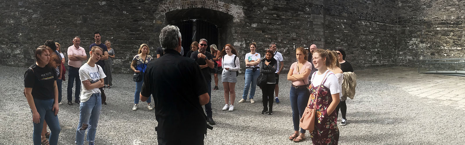 Several students look towards a man speaking whilst inside mossy, old, stone walls. 